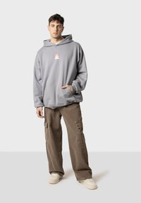 Sudadera gris con capucha y bolsillo frontal, con un gráfico de dibujos animados; combinada con pantalones cargo marrones con bolsillos laterales; deportivas blancas.
