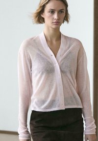Cardigan rose clair transparent à manches longues avec un décolleté en V profond, des boutons sur le devant et une coupe décontractée, associé à un pantalon foncé et ajusté.