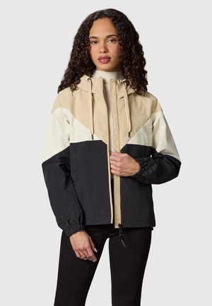 Vrouw met krullend haar, gekleed in een beige, witte en zwarte colorblock-ritsjaskap en zwarte broek, staand tegen een effen achtergrond.