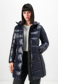 Vrouw in een glanzende donkerblauwe gewatteerde pufferjas met capuchon, witte top en zwarte broek, staand tegen een effen witte achtergrond.