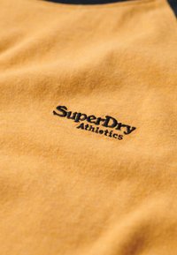 Tejido de algodón color mostaza con un logo bordado en negro de "SuperDry Athletics". Textura suave con un ligero brillo.