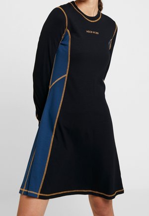 Femme portant une robe longue noire et bleue avec des coutures contrastantes orange et le logo "WOOD WOOD" sur la poitrine.