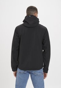 Giacca impermeabile nera con cappuccio, caratterizzata da una texture liscia, polsini regolabili e un design semplice. Indossata sopra jeans in denim blu.