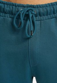 Turkise joggebukser med samlet elastisk liv, justerbar snøring og sidelommer med en glatt tekstur.