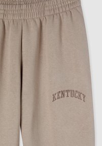 Pantaloni della tuta beige con elastico in vita e testo ricamato "KENTUCKY" in una tonalità più scura. Tessuto morbido e liscio.