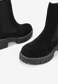 Botines de ante negro con suela de goma gruesa, paneles laterales elásticos y costuras en contraste a lo largo del borde de la suela.