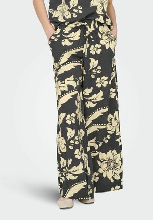 Czarne spodnie o szerokich nogawkach z kremowym kwiatowym nadrukiem, elastyczny pas, sznurek do wiązania, boczne kieszenie, noszone z beżowymi butami, dłonie w kieszeniach.