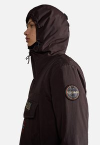 Brown hooded jacket with a textured finish, featuring a front pocket and patches on the sleeve. Drawstring detail at the hood.