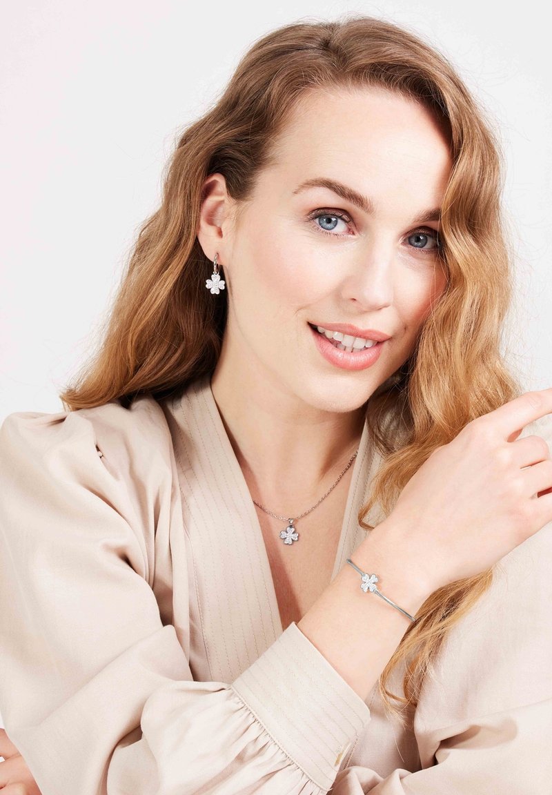 Sieradenset met een zilveren bloemenhanger ketting, bijpassende oorbellen en een armband. De bloemaccenten hebben een gestructureerde afwerking op een beige top.
