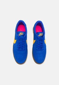 Sapatilhas de camurça azul com o logo da Nike em amarelo, apresentando biqueira arredondada, atacadores planos e forro interior rosa. Sola de borracha para tração.