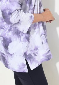 Blusa bianca e viola in stile tie-dye con maniche a tre quarti e spacchi laterali, realizzata in tessuto morbido. Abbinata a leggins scuri.