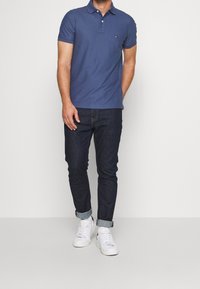 Modrá polo triko s malým logem, nošené s tmavými džíny a bílými teniskami. Krátké rukávy, klasický límec a hladká textura.