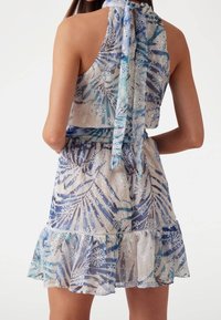 Robe légère et transparente avec un motif en dentelle florale bleu et blanc. Elle présente un col licou et un ourlet à volants superposés. Se noue dans le dos.