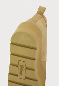 Beige zamšveida kurpes ar noapaļotu purngalu un teksturētu gumijas zoli. Izstrādājuma iekļauts vilkšanas cilpa papēžā un "Clarks Originals" zīmols uz zoles.