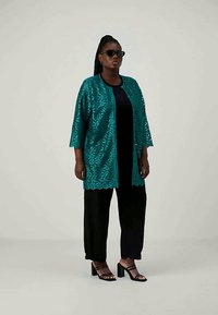 Tirkizni čipkastu kardigan s uzorkom lista, s rukavima do tri četvrtine, nosi se preko crne majice i crnih hlača, kombiniran s crnim sandalama.