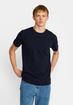 Jeune homme aux cheveux courts portant un t-shirt bleu marine à col rond avec une poche poitrine et un jean bleu, debout devant un fond uni.