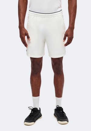 Homme portant un short de sport blanc, des chaussettes blanches, des chaussures de sport noires et un haut blanc, debout devant un fond uni.