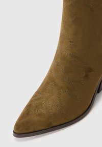 Bota de tobillo de ante marrón con puntera afilada, que presenta patrones de costura decorativos y un tacón bajo. Textura suave con un ligero brillo.