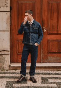 Modra jeans jakna z dvema žepoma na prsih, v kombinaciji z usklajenimi jeans hlačami. Rjavi čevlji z vezalkami, ki stojijo ob lesenih vratih.