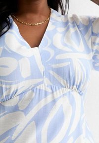 Vaaleansininen mekko, jossa on hulmuava muotoilu ja valkoisia abstrakteja kuvioita. Kangas on pehmeä ja teksturoitu, jossa on lovekaulus ja kerrottu vyötärö.