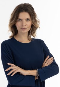 Navyblaues Langarmshirt mit einem runden Halsausschnitt. Die Arme des Modells sind verschränkt, wobei am Handgelenk ein Armband aus verschiedenen Texturen und Farben zu sehen ist.