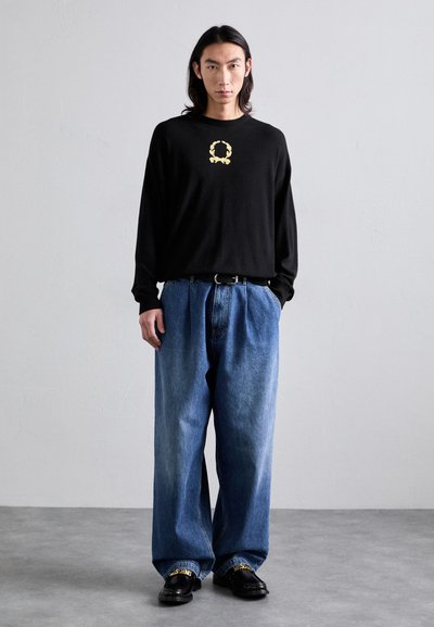 Schwarzes Langarmshirt mit goldener Kranz-Logo; lockere blaue Jeans; schwarze Schuhe mit goldenen Akzenten; glatte Texturen.