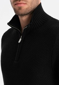 Maglione nero in maglia texture con colletto alto e zipper a metà. Primo piano che mostra la trama voluminosa e il dettaglio della zip argentata.