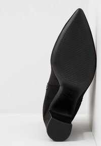 Zapato de tacón alto de ante negro con punta afilada. Cuenta con una suela texturizada y un tacón liso y curvado. Detalle de cremallera en el costado.