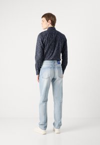 Camicia a maniche lunghe con motivo floreale blu, abbinata a jeans azzurri. I jeans hanno tasche posteriori visibili e una patch con logo blu.