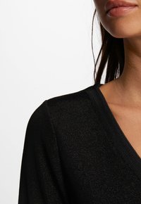 Schwarzes V-Ausschnitt-Top mit funkelnder Textur, gerippten Kanten und taillierter Passform. Der Stoff zeigt einen subtilen Schimmer auf einem hellen Hintergrund.