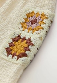 Cremefarbener Stoff mit zwei mehrfarbigen Häkelquadraten in Gelb, Braun, Pink und Weiß.
