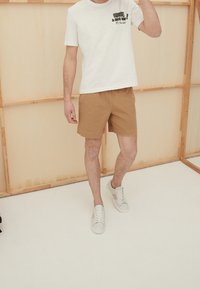 Camiseta blanca de manga corta con estampado negro, combinada con pantalones cortos beige y zapatillas grises. Atuendo casual en un modelo de pie contra un fondo claro.