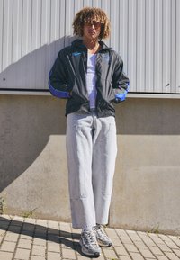 Ung person med krøllet hår iført orange farvede solbriller, sort og blå Nike-jakke, hvid tanktop, lyse brede jeans og sneakers, som står udenfor.