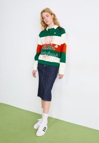 Polo de manga comprida às riscas verdes, vermelhas e brancas com logótipo e texto, combinada com uma saia de ganga escura até meio da perna e ténis brancos.