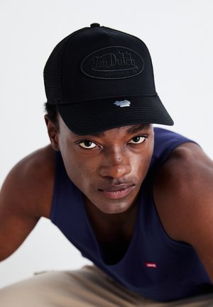 Jeune homme portant une casquette noire Von Dutch et un débardeur bleu marine, penché en avant et regardant directement la caméra.