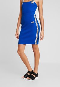 Modré přiléhavé šaty s nastavitelnými ramínky, s černobílým logem Adidas po stranách. Černé sportovní sandály s bílými podrážkami.