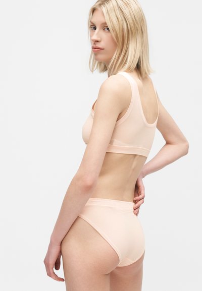 Γυναίκα με ξανθά μαλλιά φοράει ελαφρώς ροδακινί χρωματιστό σουτιέν και εσώρουχα, ποζάροντας σε πλάγια όψη με το ένα χέρι στη μέση, μπροστά από ένα απλό φόντο.