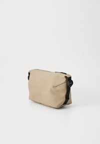 Beige vanntett pose med en myk tekstur, rektangulær form, topplukking med glidelås og justerbare svarte stropper på sidene.