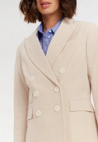Blazer double boutonnage beige avec des rayures subtiles. Il dispose de grands boutons blancs, de revers crantés et d'un tissu texturé. Des poches latérales sont incluses.
