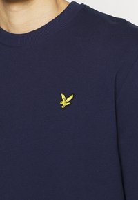 Sudadera azul marino de tejido suave con un cuello redondeado acanalado y un pequeño logo de pájaro amarillo bordado en el lado izquierdo del pecho.