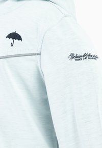 Top in tessuto azzurro chiaro con un design di ombrello nero ricamato e testo sulla manica. Texture liscia con una leggera lucentezza.