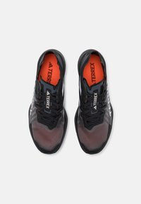 Black athletic shoes with mesh upper, textured pattern, reflective accents, and orange insole. Features logo and traditional lace-up design.