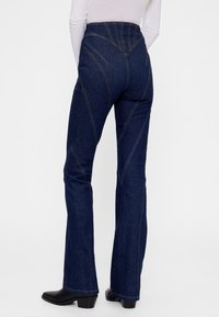 Sötétkék, magas derekú, bővülő szárú farmernadrág, látható, kontrasztos varrással és ívelt varrási részletekkel. Simaságú anyag, testhezálló kialakítás.