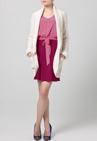 Neulepaita vaaleassa sävyssä, kerroksittain vaaleanpunaisen vyötärösidoksella varustetun mekon päällä. Mekossa on kellotettu helma ja siniset korkeakorkoiset kengät. Tasaiset rakenne-ilmeet ovat ilmeisiä.