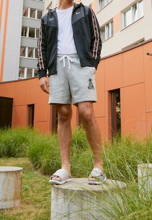 Persona con chaqueta negra de Nike, camiseta blanca, pantalones cortos grises de Nike y sandalias desparejadas de tipo deslizador, de pie sobre un bloque de concreto al aire libre.