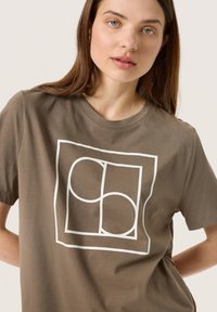 Brun bomull t-shirt med en vit geometrisk grafisk design på framsidan. Rundad halsringning och korta ärmar. Slät yta.