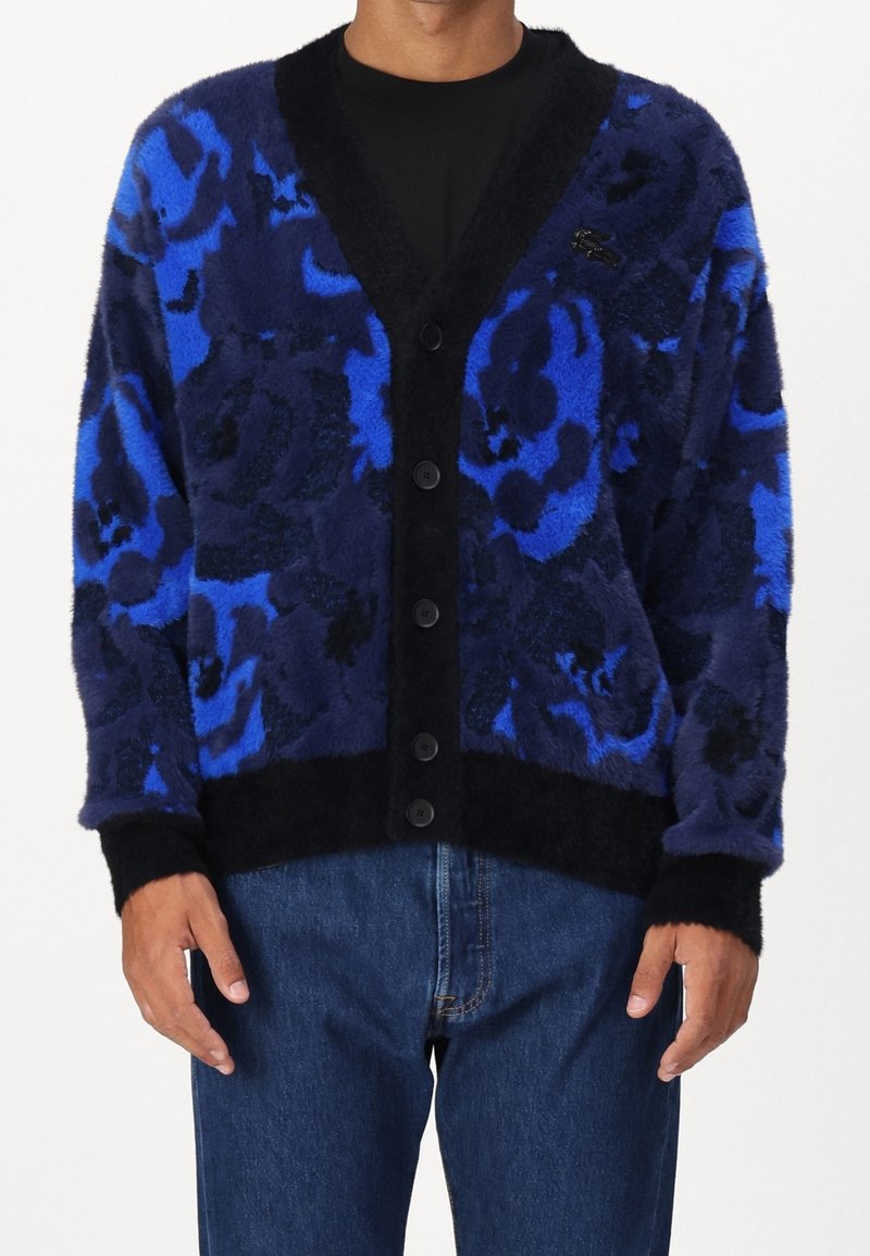 Homme portant un cardigan duveteux à motifs bleus et noirs avec des boutons noirs par-dessus une chemise noire et un jean bleu.