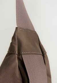 Brun taske med et struktureret lilla strop. Stoffet har glat, holdbart nylon med synlige sømme og en polstret fastgørelsesdetalje.