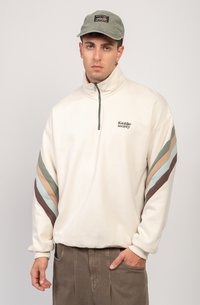 Pullover in pile di colore crema con zip verde, caratterizzato da strisce diagonali in marrone, azzurro chiaro e tonalità attenuate. Logo sul petto.