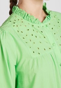 Blusa verde chiaro con scollatura a volant, caratterizzata da un design in pizzo floreale e chiusura a bottoni. Materiale in cotone morbido con dettagli arricciati.